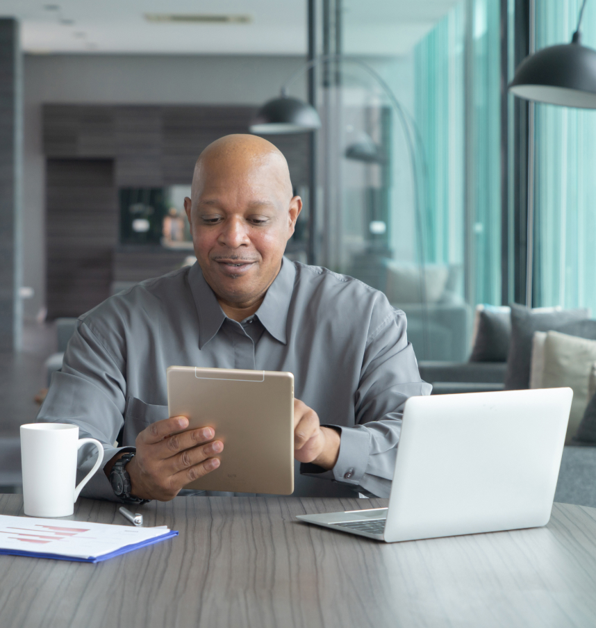 Homem sentado em frente a uma mesa mexendo em um tablet com um notebook em sua frente.